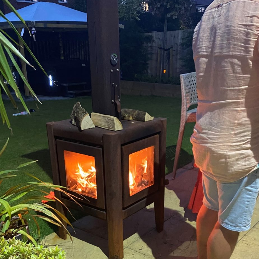 L'Bode Cube Corten Edition Wood Burner with logs on top in a garden setting