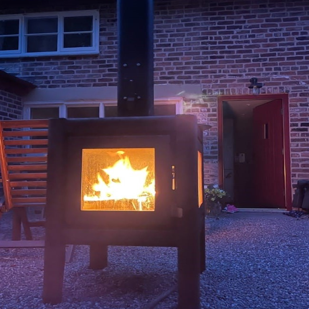 L'Bode Cube Corten Edition Wood Burner outdoors with firepit lit